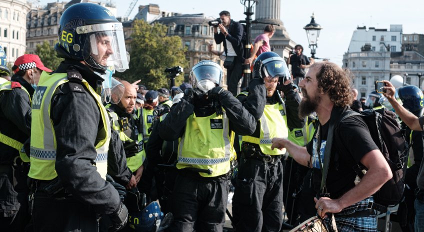 Polizeibeamte in Schutzkleidung und gelben Westen stehen einer Gruppe von Demonstranten gegenüber. Einige Demonstranten filmen die Szene, während andere diskutieren. Im Hintergrund sind städtische Gebäude und ein Denkmal erkennbar.